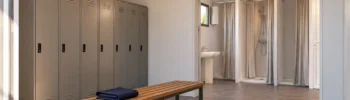 Interior ilustrativo de un vestuario prefabricado con taquillas, banco de madera y una zona separada con duchas y lavabo.