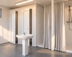 Interior de un módulo sanitario con cabinas de aseo, lavabo central y duchas separadas por cortinas.