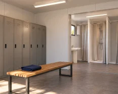 Interior ilustrativo de un vestuario prefabricado con taquillas, banco de madera y una zona separada con duchas y lavabo.
